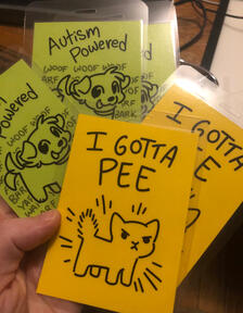Mini badges to wear at cons that have an annoyed-looking cat and the caption I GOTTA PEE and a happy little dog with the caption Autism Powered Bark Machine and barking in the background! They are on fluorescent yellow and green cardstock respectively.