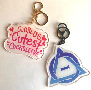 Two acrylic 4” inch charms on a white background. One has gold hardware and features the text “World’s Cutest Cocksleeve” in hot pink with hearts around it on a white background. The other has black hardware and features a blue/violet therian symbol.