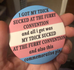 A photo of a hand holding an oversized button pin. The background is a trans flag. Black letters read: “I GOT MY TDICK SUCKED AT THE FURRY CONVENTION and all i got was MY TDICK SUCKED AT THE FURRY CONVENTION and also this commemorative pin”