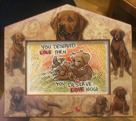 A decorative frame featuring watercolor chocolate labrador retriever puppies and adults with a watercolor painting inside it of an identical adult and puppy labrador laying happily together in a field with a red ball, wagging their tails as they touch nose
