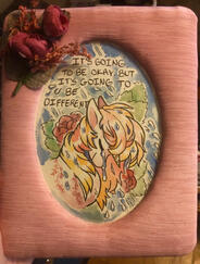 It's Going to be Okay, But It's Going to Be Different A watercolor painting in a pink fabric frame with faux roses attached to the edge. A unicorn in shades of pink, gold and orange smiles gently while openly weeping. Roses bloom behind them. Rain pours and tears fall. A caption reads: "IT'S GOING TO BE OKAY,